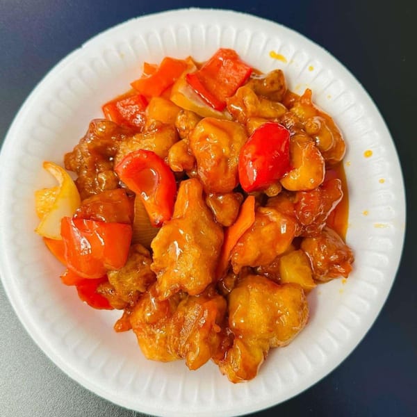 Sweet and sour chicken with red and yellow bell peppers in a white paper bowl.