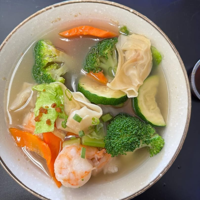 Wonton soup with shrimp, broccoli, carrots, zucchini, and green onions in a white bowl.